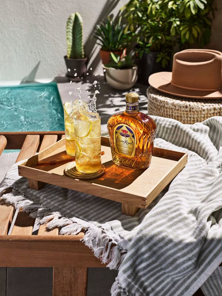 A wooden tray holding a glass of lemon-infused drink and a bottle of Crown Royal whiskey, surrounded by potted plants and cozy textiles.