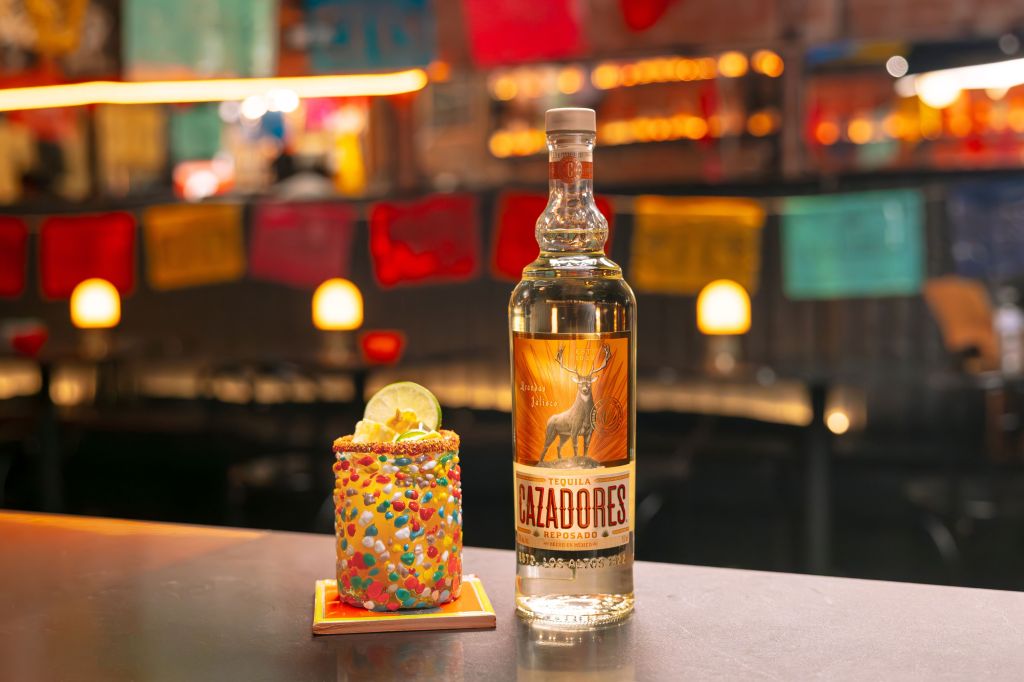 A bottle of Cazadores tequila and a colorful cocktail with sprinkles on a bar with festive lighting in the background.