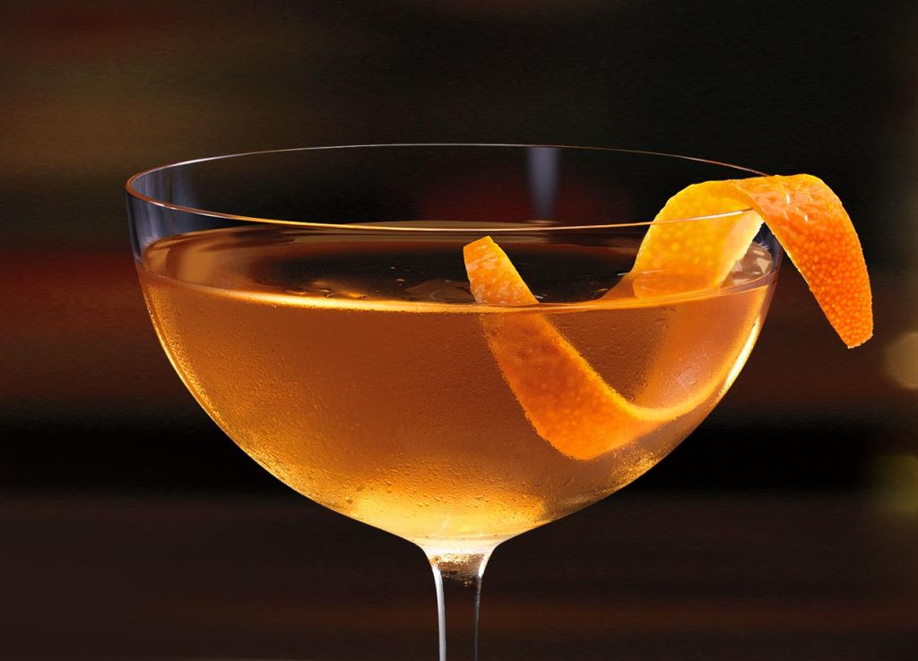 A glass of amber-colored cocktail garnished with an orange twist, set against a dark background.