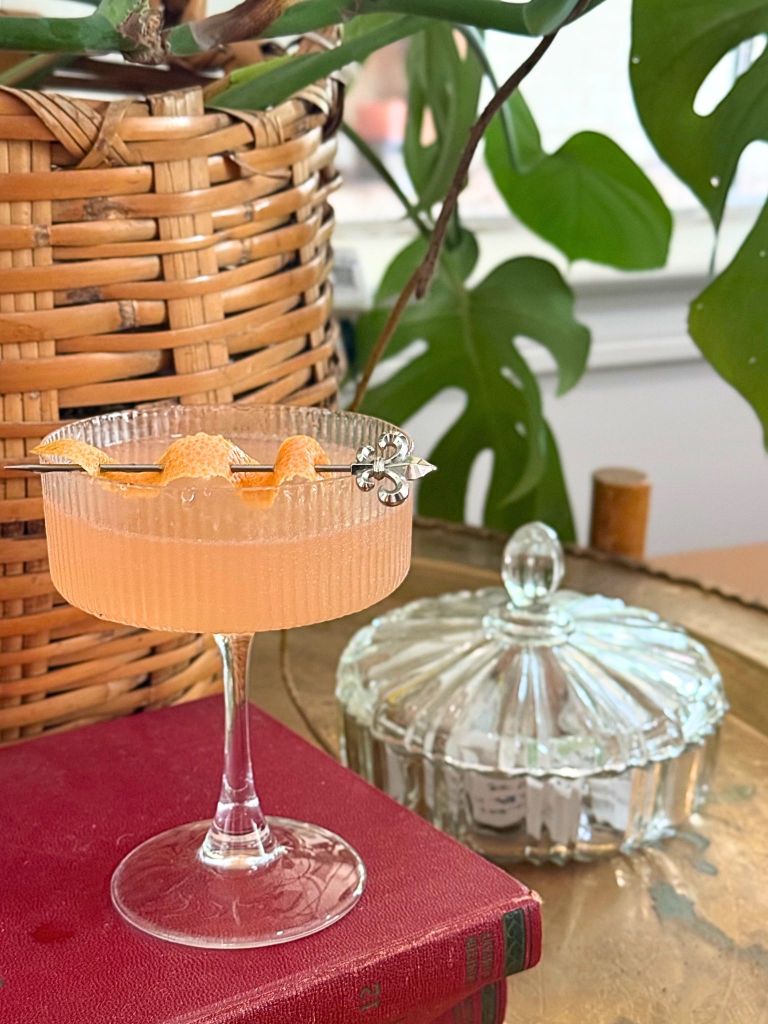 A glass of orange cocktail garnished with orange slices, sitting on a red book in front of a woven basket and lush green leaves.