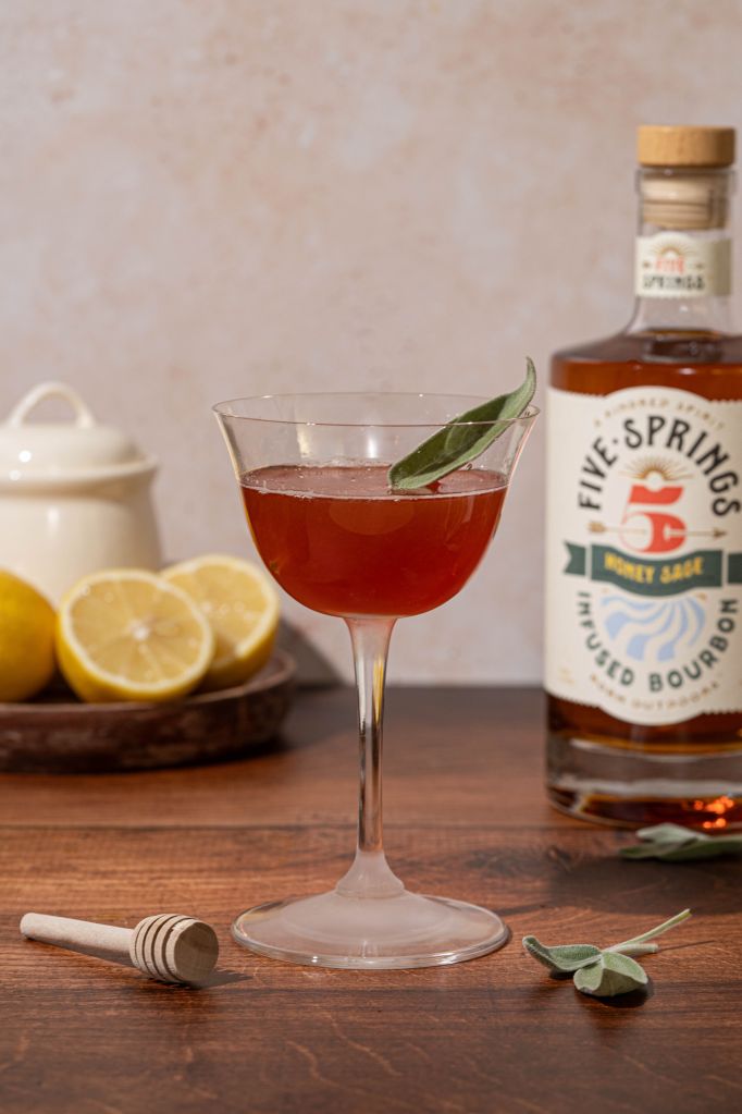 A glass of amber-colored cocktail garnished with a sage leaf, accompanied by a bottle of Five Springs Bourbon and lemon slices on a wooden table.