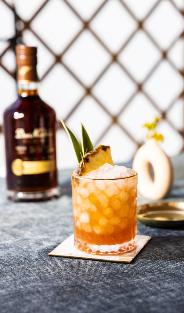 A glass of orange cocktail with pineapple garnish, set against a blurred diamond-patterned background and a bottle of dark liquor.