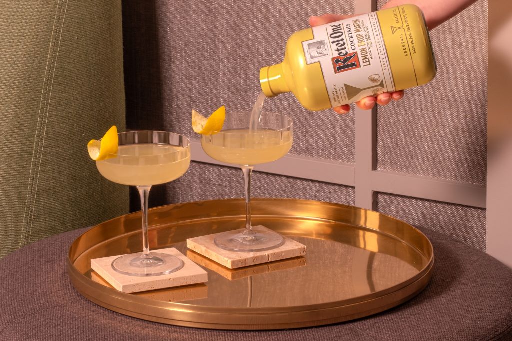 A person pouring a yellow liquid from a bottle into a glass on a gold tray, with sliced citrus garnishes.