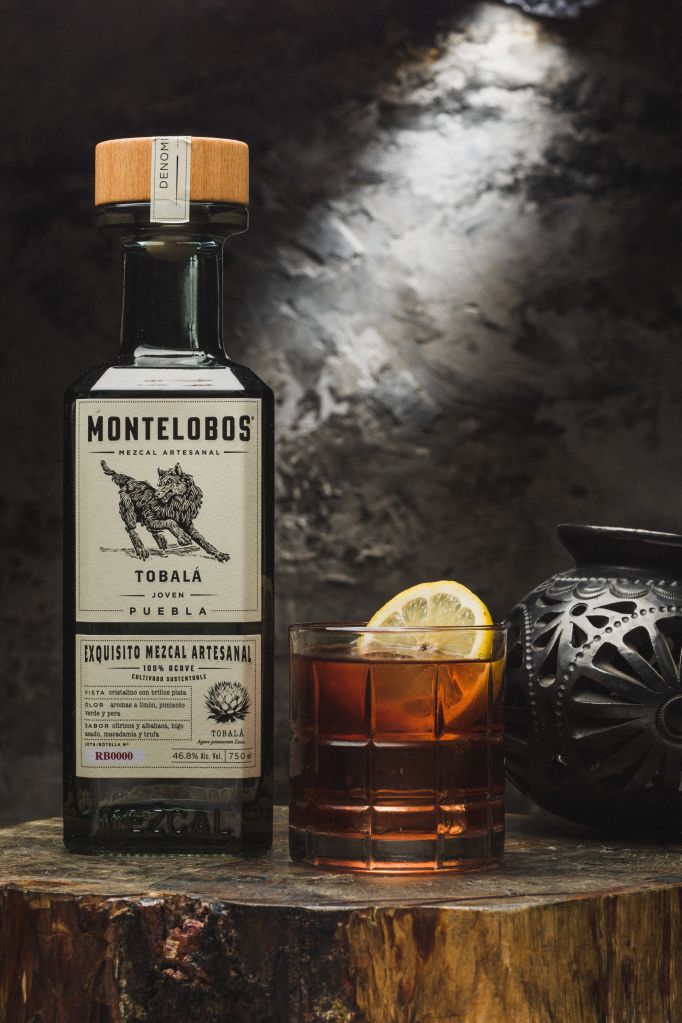 A bottle of Montelobos Mezcal Artesanal Tobala Joven Pueblo and a glass of the mezcal on a wooden surface, with a lemon slice and a decorative skull-patterned glass.
