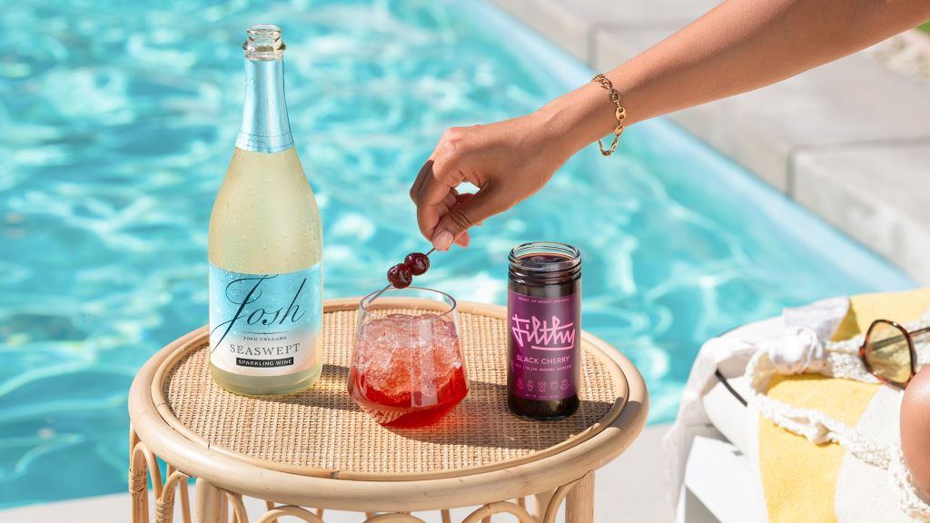 A hand holding a glass of red cocktail on a tray with a bottle of Josh Seawept sparkling wine and a jar of black cherry jam, set against a swimming pool.
