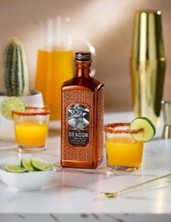 A bottle of Malfy gin surrounded by orange slices, a glass of orange cocktail, and a cactus plant on a marble surface.