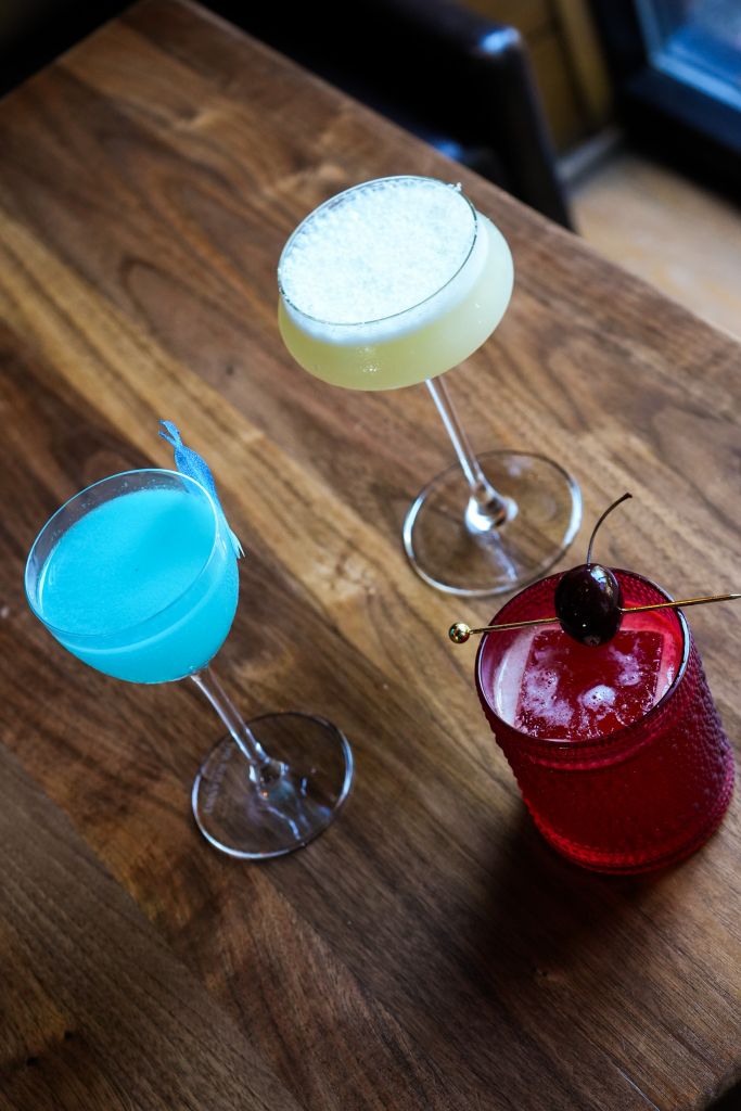 Three cocktail glasses on a wooden table, containing a blue drink, a yellow drink with a white frothy top, and a red drink with a cherry garnish.