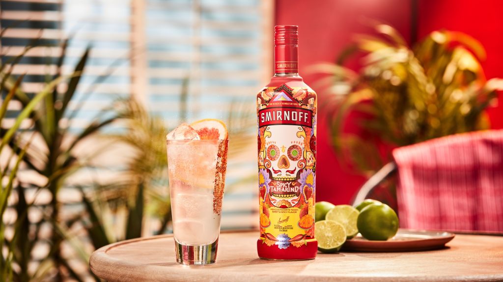A red Smirnoff vodka bottle and a cocktail glass with a lime wedge on a wooden table, surrounded by tropical foliage.
