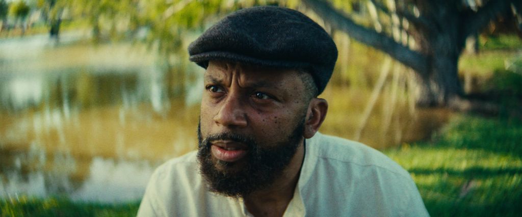 A bearded man wearing a black cap and a white shirt, standing in a lush, green outdoor setting.