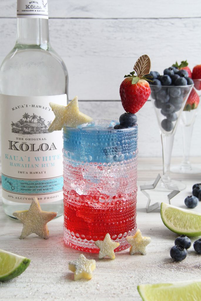 A colorful cocktail glass filled with a layered red and blue drink, garnished with a strawberry and blueberries. A bottle of Koloa Hawaiian rum is visible in the background.