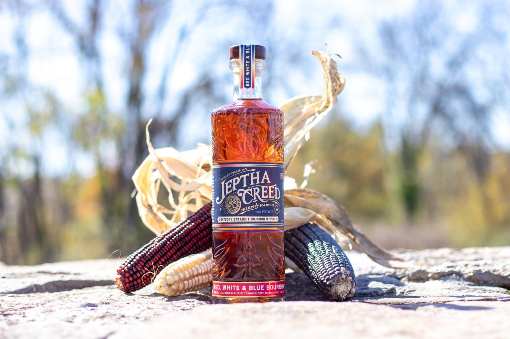 A bottle of Jeptha Creed whiskey surrounded by autumn leaves and corn cobs on a wooden surface.