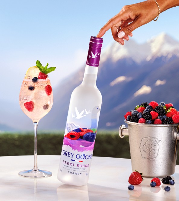 A hand holding a bottle of Grey Goose Berry Rouge vodka, with a glass of mixed berries and a mountain landscape in the background.