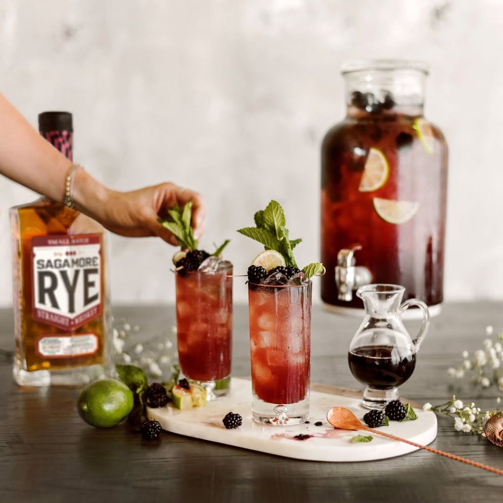 A glass of red cocktail garnished with blackberries, mint, and lemon slices, alongside a bottle of Sagamore Rye whiskey and other cocktail ingredients on a dark surface.