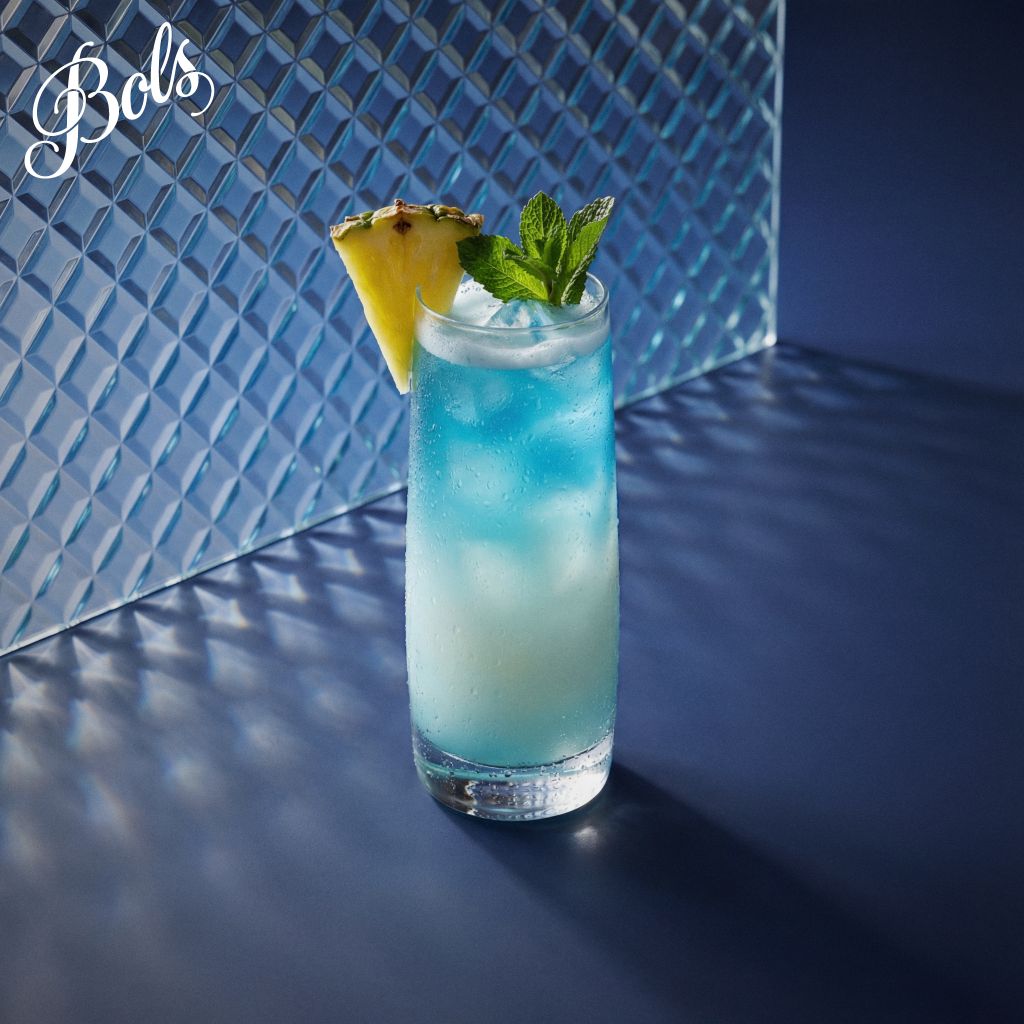 A glass filled with a blue-colored beverage, garnished with a pineapple slice and mint leaves, set against a geometric blue background.
