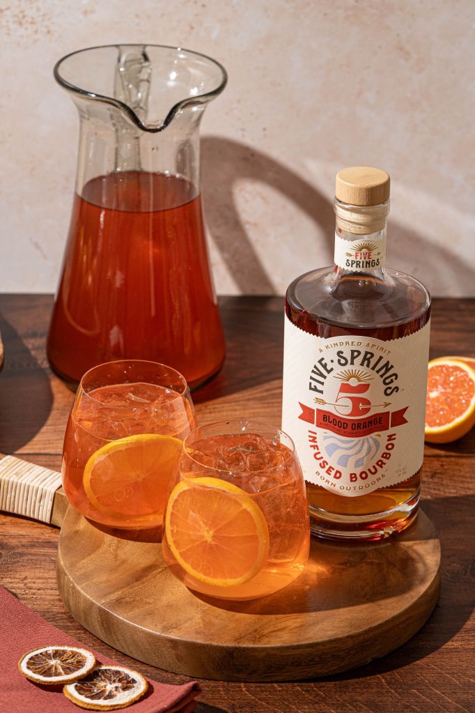 A glass pitcher filled with an orange-colored beverage, accompanied by a bottle of Five Springs Blood Orange liquor and glasses with orange slices on a wooden surface.