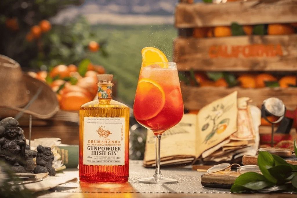 A glass of orange cocktail with a lemon slice, next to a bottle of Drumshando Gunpowder Irish Gin, surrounded by citrus fruits and other decorative elements.