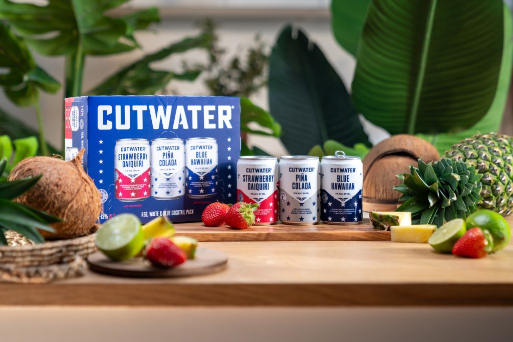 Assortment of Cutwater canned cocktails including Strawberry Daiquiri, Pina Colada, and Blue Hawaiian, surrounded by tropical fruits and greenery.