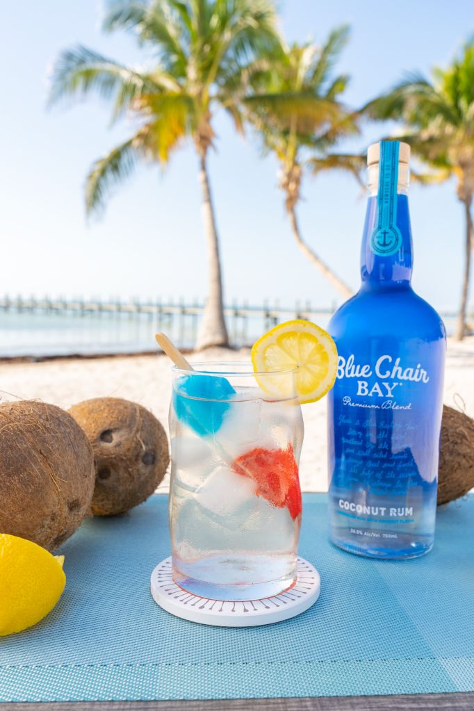 A tropical beach scene with palm trees, a blue bottle of Blue Chair Bay coconut rum, and a glass of a colorful cocktail garnished with fruit.