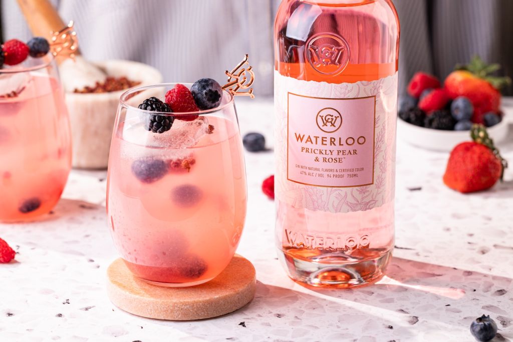 A glass of pink cocktail garnished with fresh berries, alongside a bottle of Waterloo Prickly Pear & Rose gin.