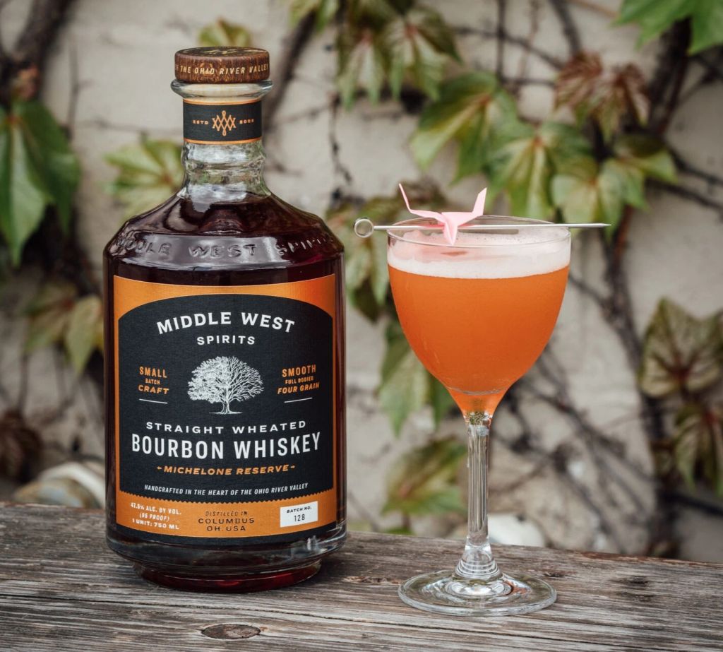 A bottle of Middle West Spirits Straight Wheated Bourbon Whiskey and a glass of an orange cocktail on a wooden surface with greenery in the background.