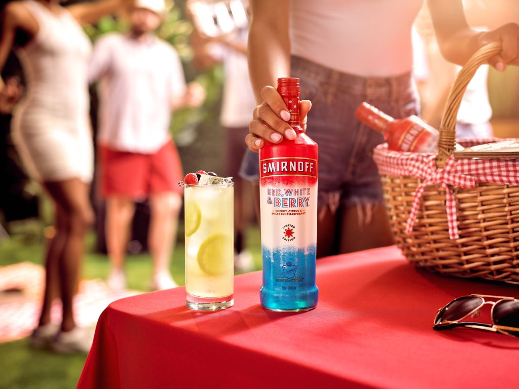 A person holding a bottle of Smirnoff Red, White & Berry vodka and a glass of lemonade on a red tablecloth with a picnic basket in the background.