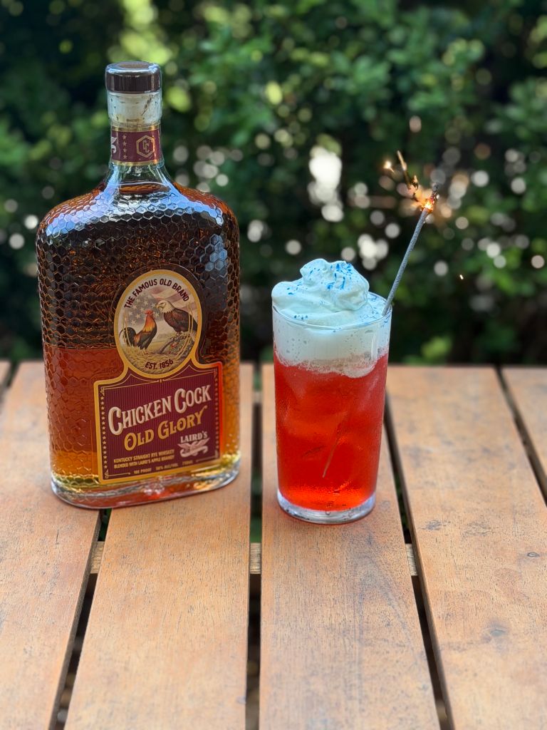 A bottle of Chicken Cock Old Glory Kentucky straight rye whiskey and a glass of red cocktail with whipped cream and a sparkler on a wooden table against a blurred green background.