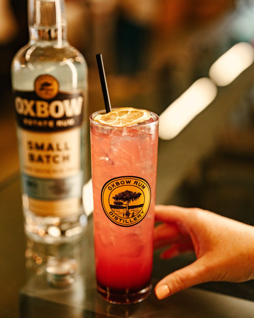 A glass of pink cocktail with a lemon slice, served with a bottle of Oxbow rum on a wooden surface.
