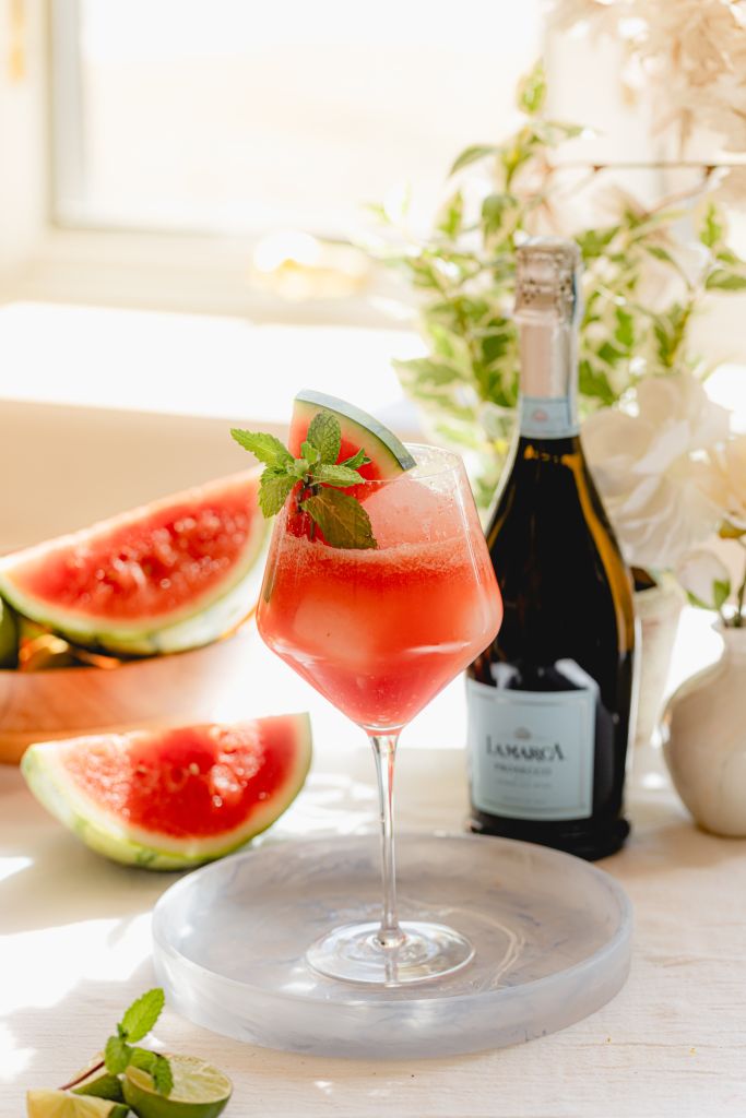 A glass of red watermelon cocktail garnished with mint, accompanied by a bottle of Lamarca wine and sliced watermelon on a table.