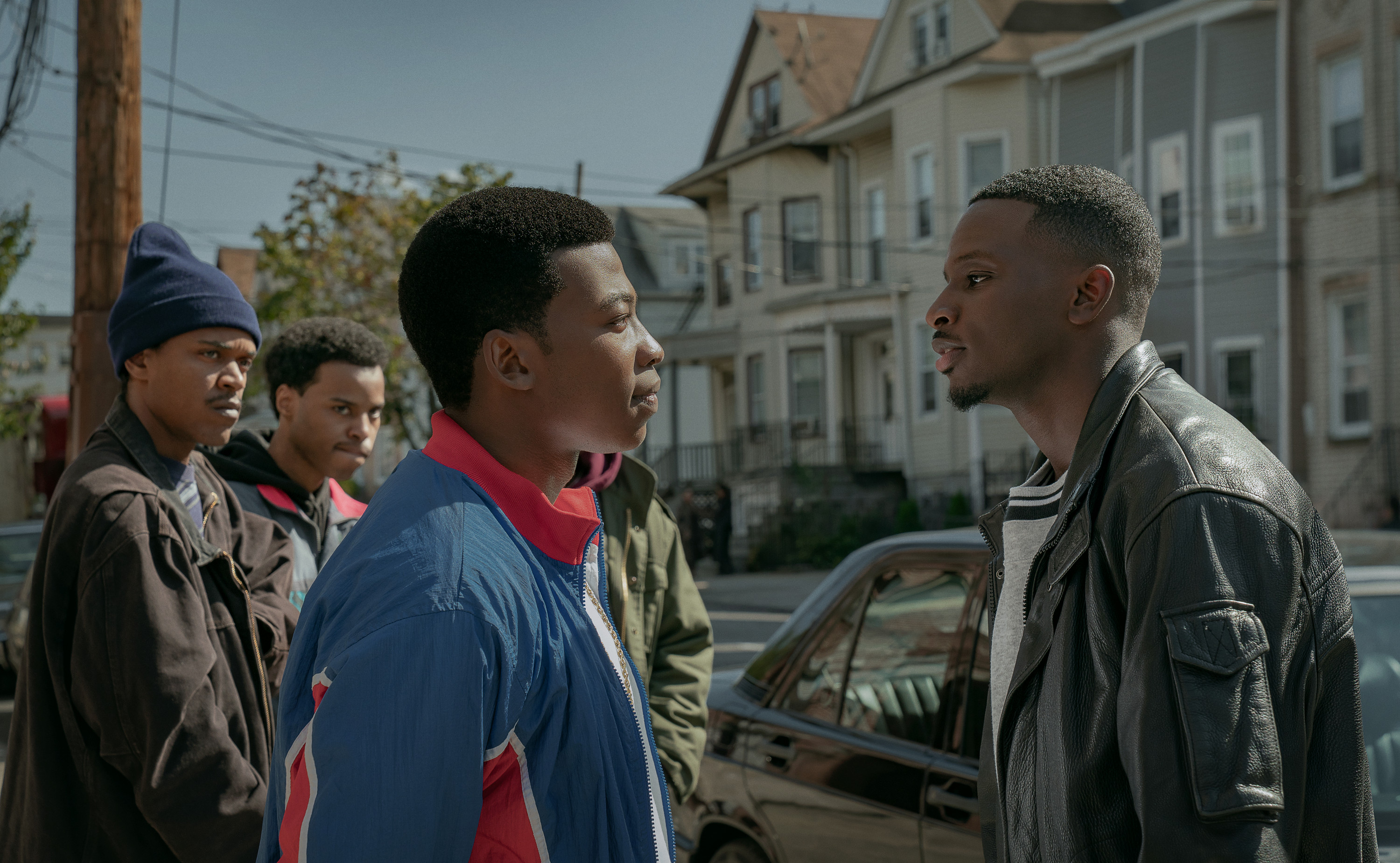 Four young Black men standing on a city street, one wearing a blue and red jacket, another a leather jacket, facing each other in a tense confrontation.