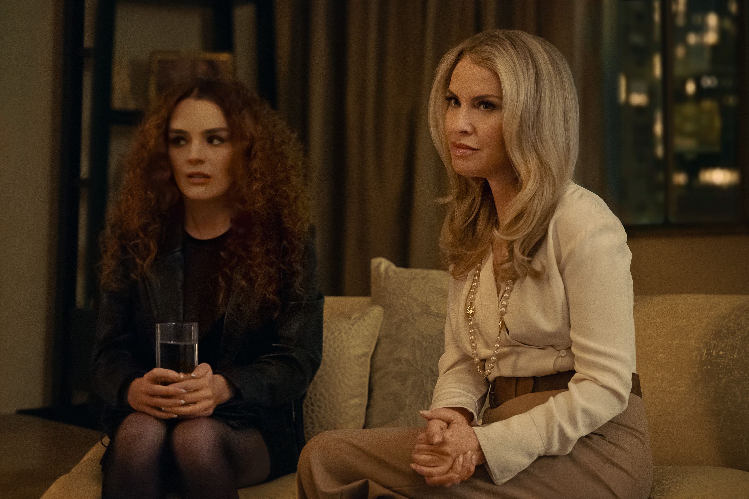 Two women, one with curly red hair and one with long blonde hair, sitting on a couch in a dimly lit room. The blonde woman is wearing a beige blouse and the brunette woman is holding a glass.