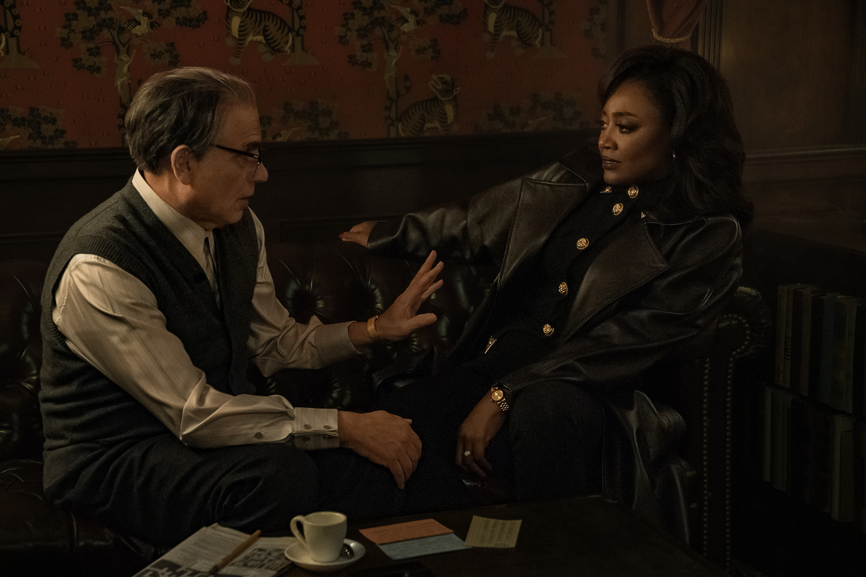 Two people, a man and a woman, sitting and conversing in a dimly lit room with decorative wall hangings.