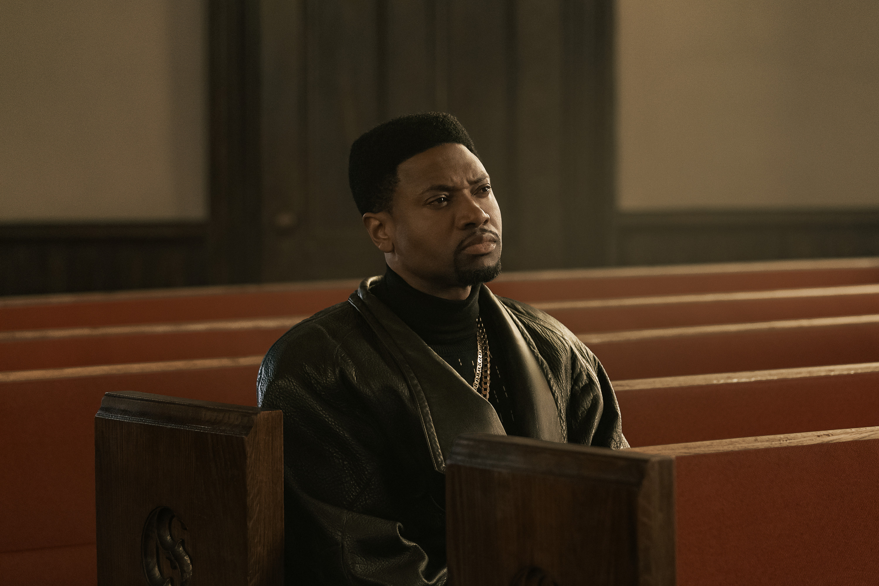 A serious-looking man wearing a black jacket and necklace, sitting alone in a church pew.