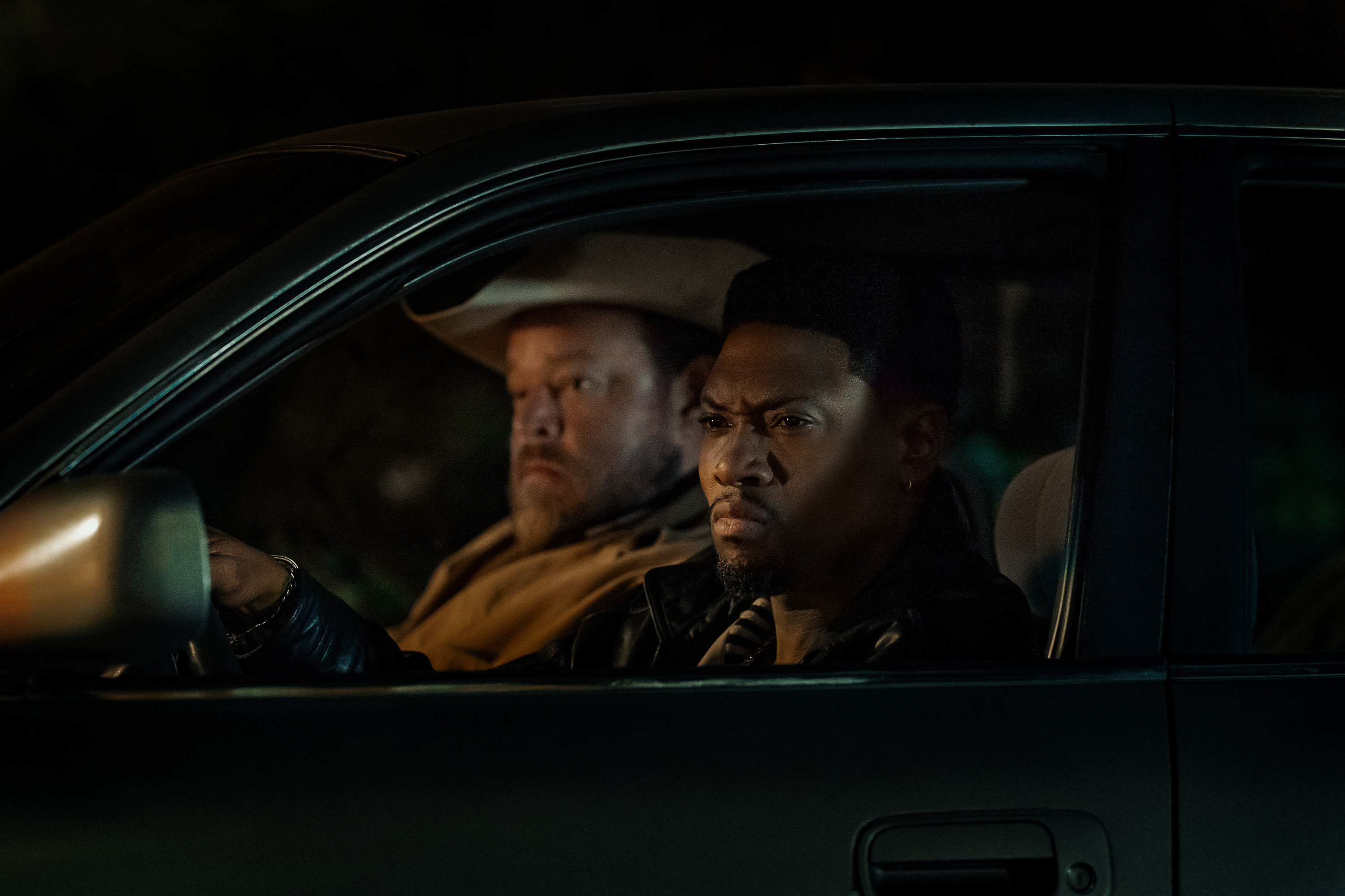 Two men sitting in a dark car, one with a serious expression and the other with a concerned look on his face.