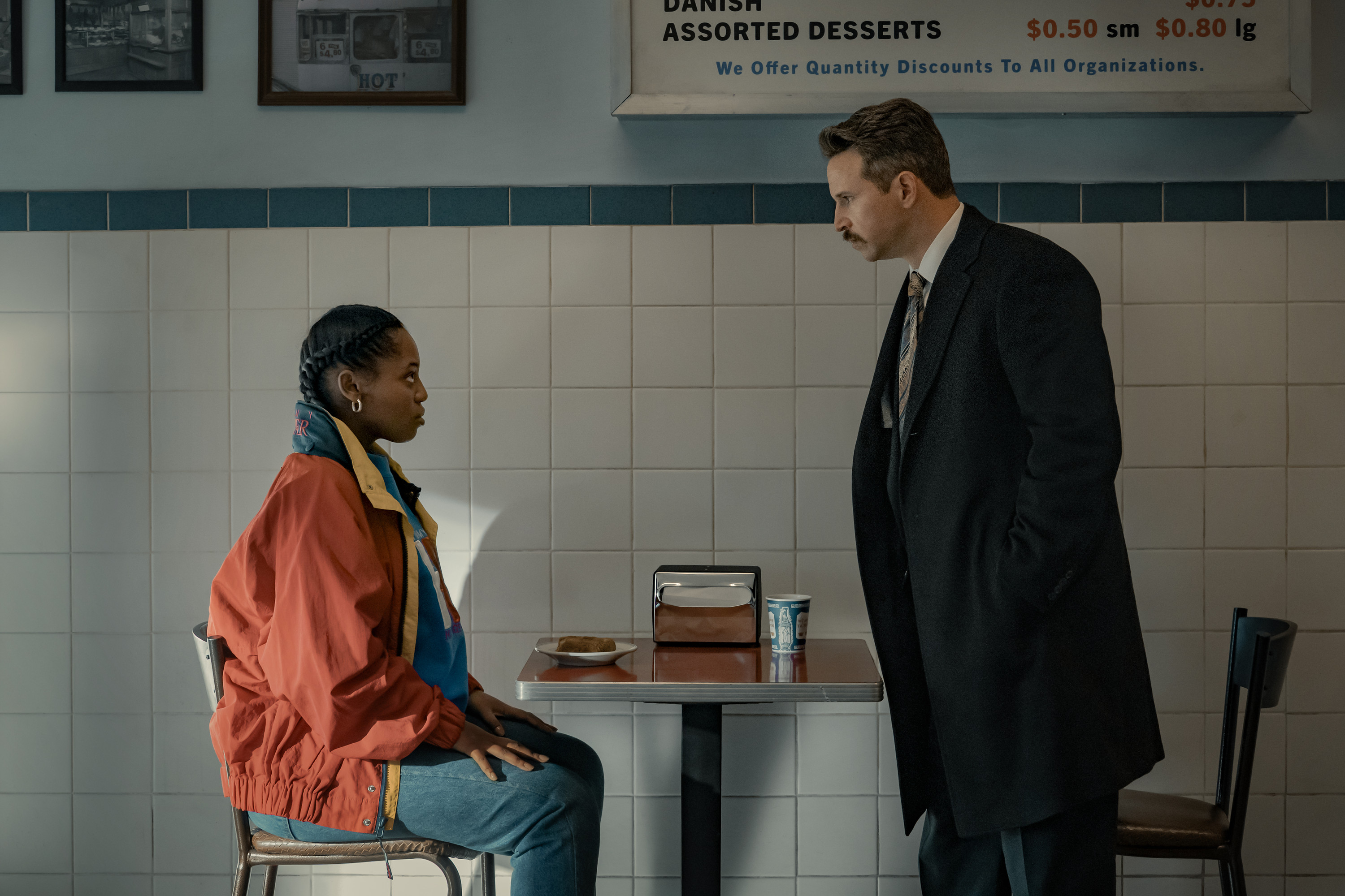 Two people, a man in a suit and a woman in a colorful jacket, standing in a tiled room with a sign for "Danish Assorted Desserts" visible.