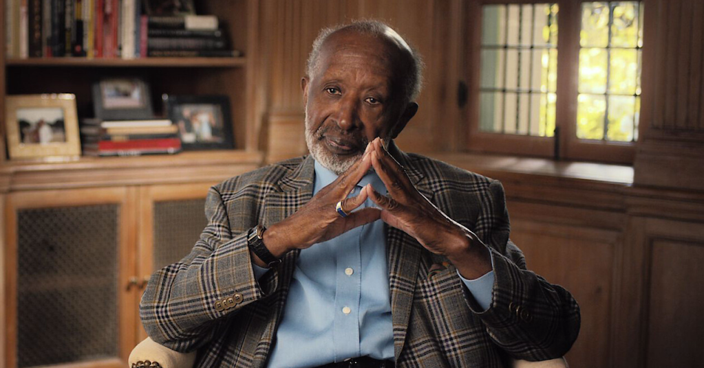 An elderly African American man in a plaid jacket sitting in a wooden-paneled room, his hands clasped in a thoughtful gesture.