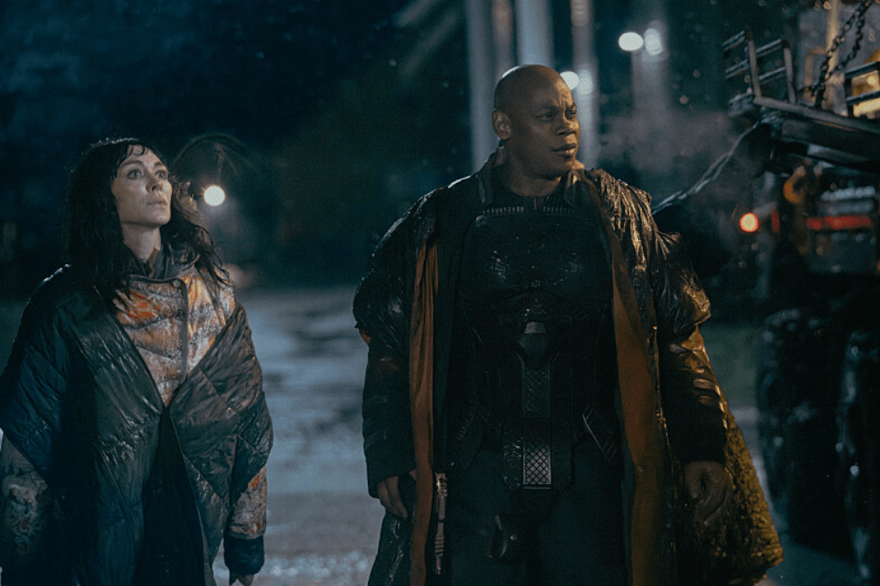 Two people, a man and a woman, walking together at night in a snowy, wooded setting.