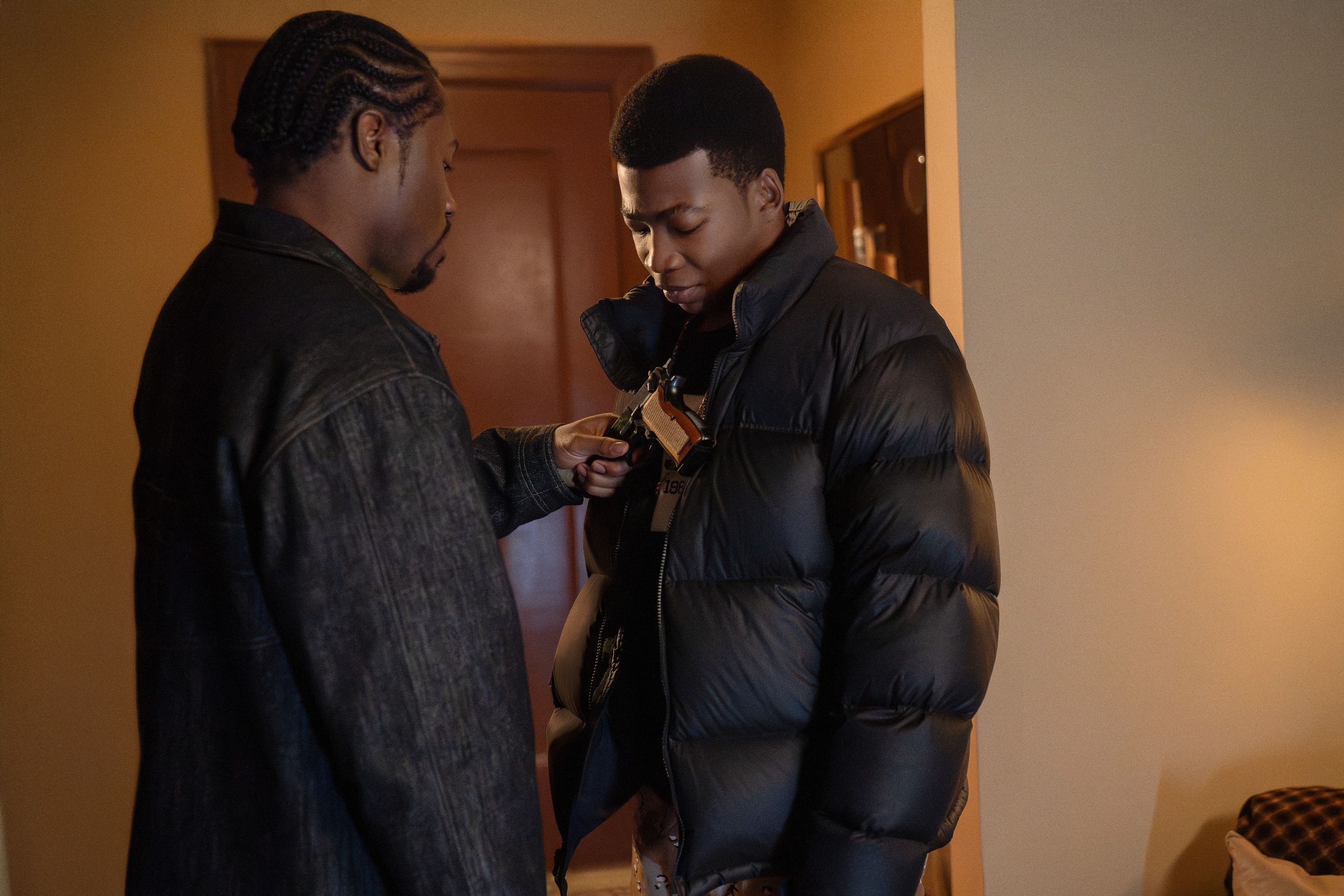 Two young Black men in winter coats standing in a doorway, one holding a small object in his hand.