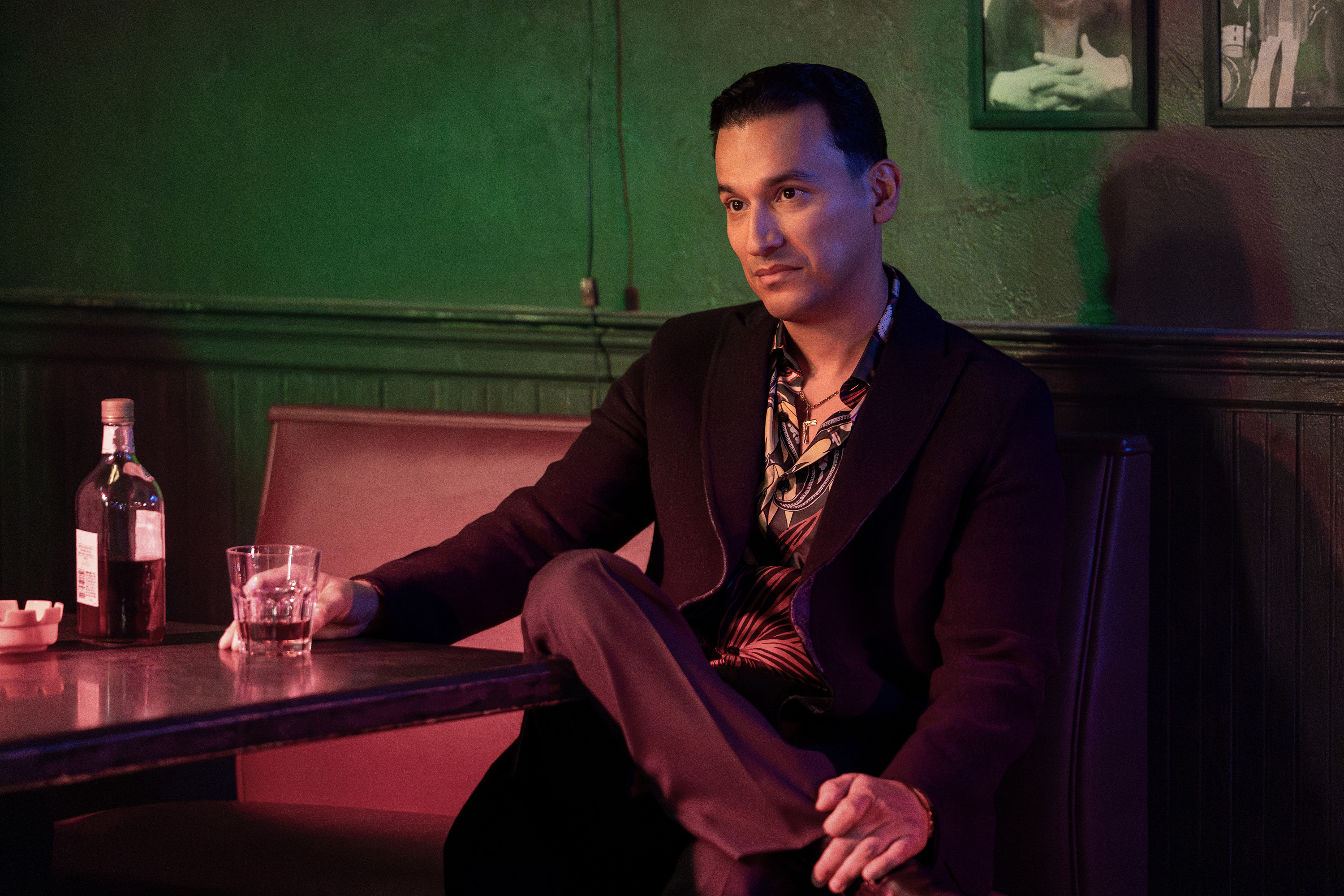 A man in a dark suit sitting at a bar, holding a glass of liquor and looking pensive in a dimly lit room with green walls and framed artwork.