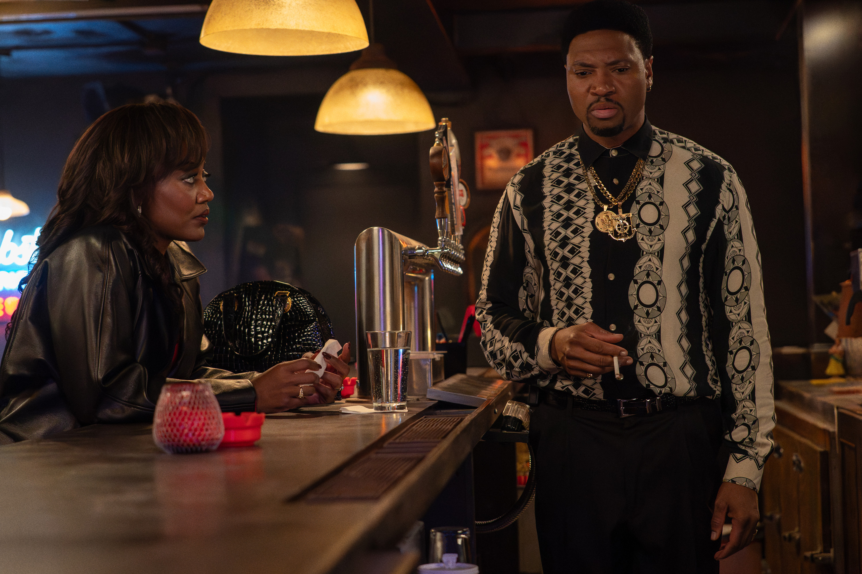 Two people, a woman and a man, standing at a bar counter in a dimly lit room. The man is wearing an ornate patterned jacket and necklace.
