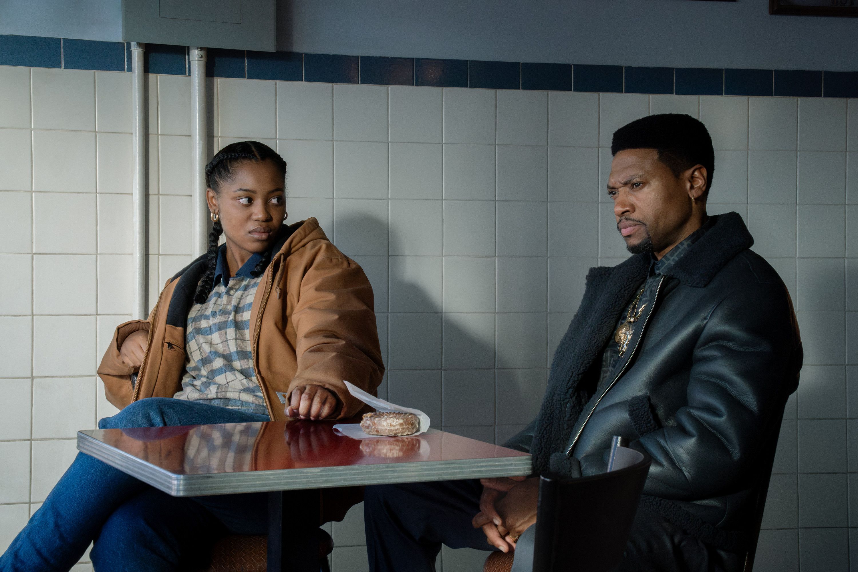 Two people, a man and a woman, sitting at a table in what appears to be a diner or cafe. The man is wearing a dark jacket and the woman is wearing a brown coat.