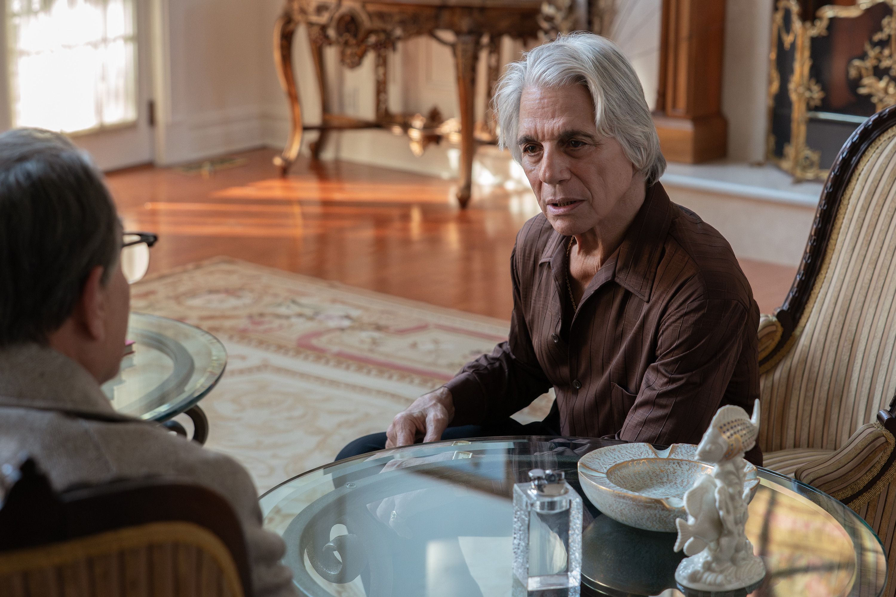 An elderly man with gray hair sitting at a table in an ornate room, looking at a younger man across from him.