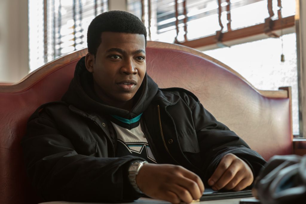 A young Black man sitting in a chair, wearing a black jacket and looking directly at the camera with a serious expression.
