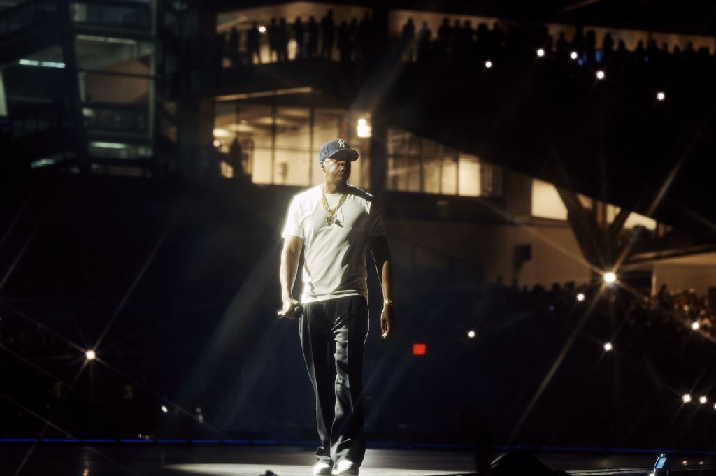 A person wearing a white t-shirt, baseball cap, and necklace standing on a stage at night with lights and buildings in the background.