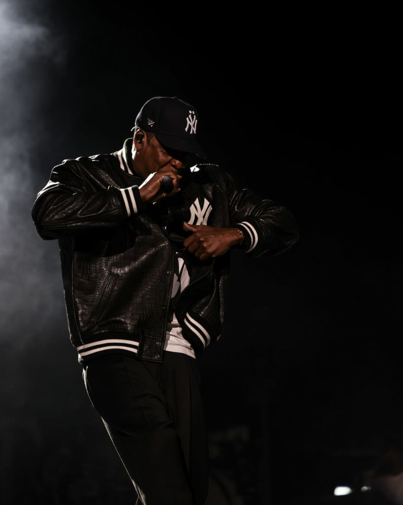 A person wearing a New York Yankees baseball cap and jacket performing on stage in a dark, smoky setting.