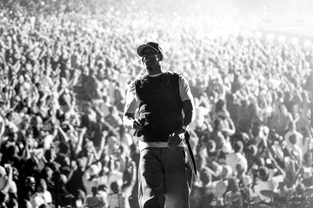 A performer on stage in front of a large crowd, wearing a black hat and shirt.