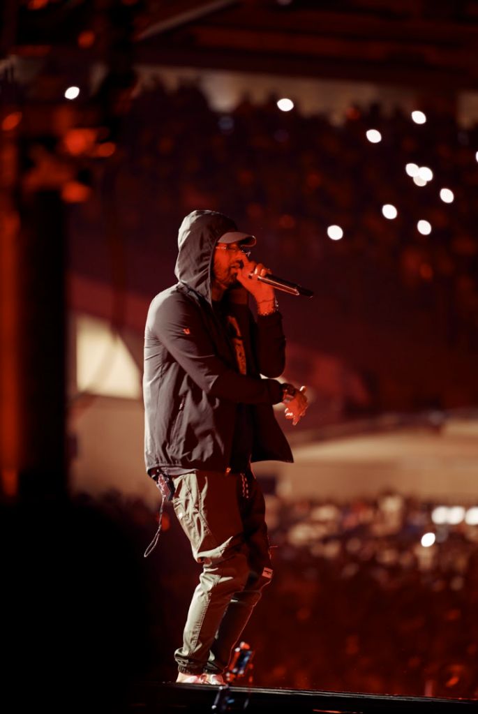 A person in a hooded jacket performing on a stage at night, with colorful lights in the background.