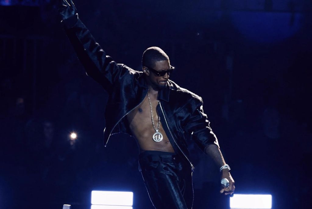 A man wearing sunglasses and a black jacket performing on stage in a dimly lit setting.