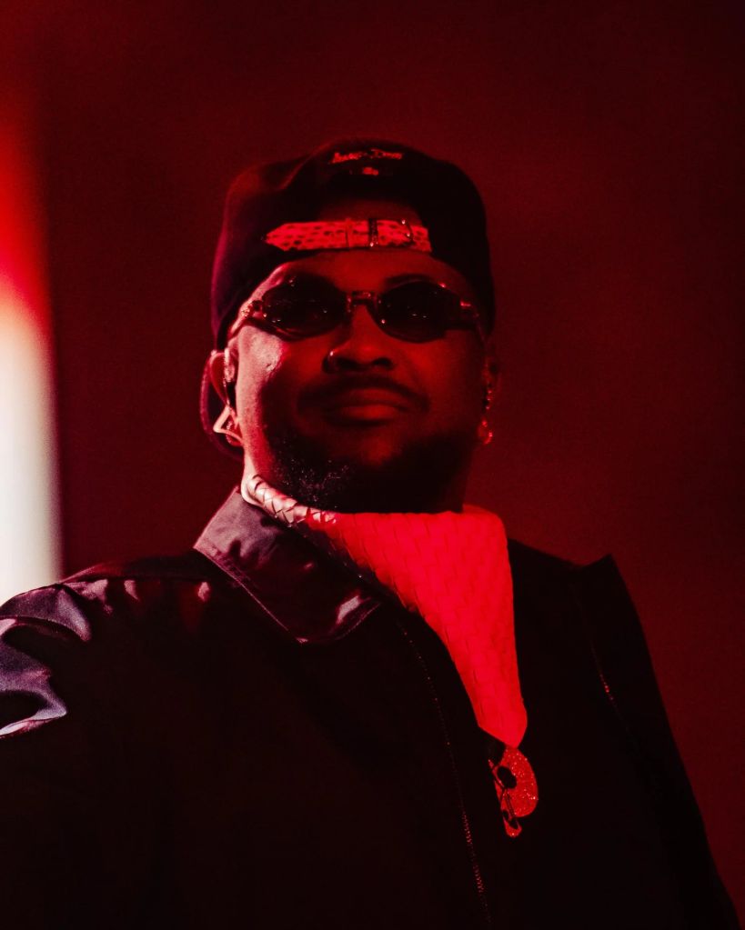 A person wearing a black hat and sunglasses, with a red scarf, standing in a dimly lit setting with a red background.