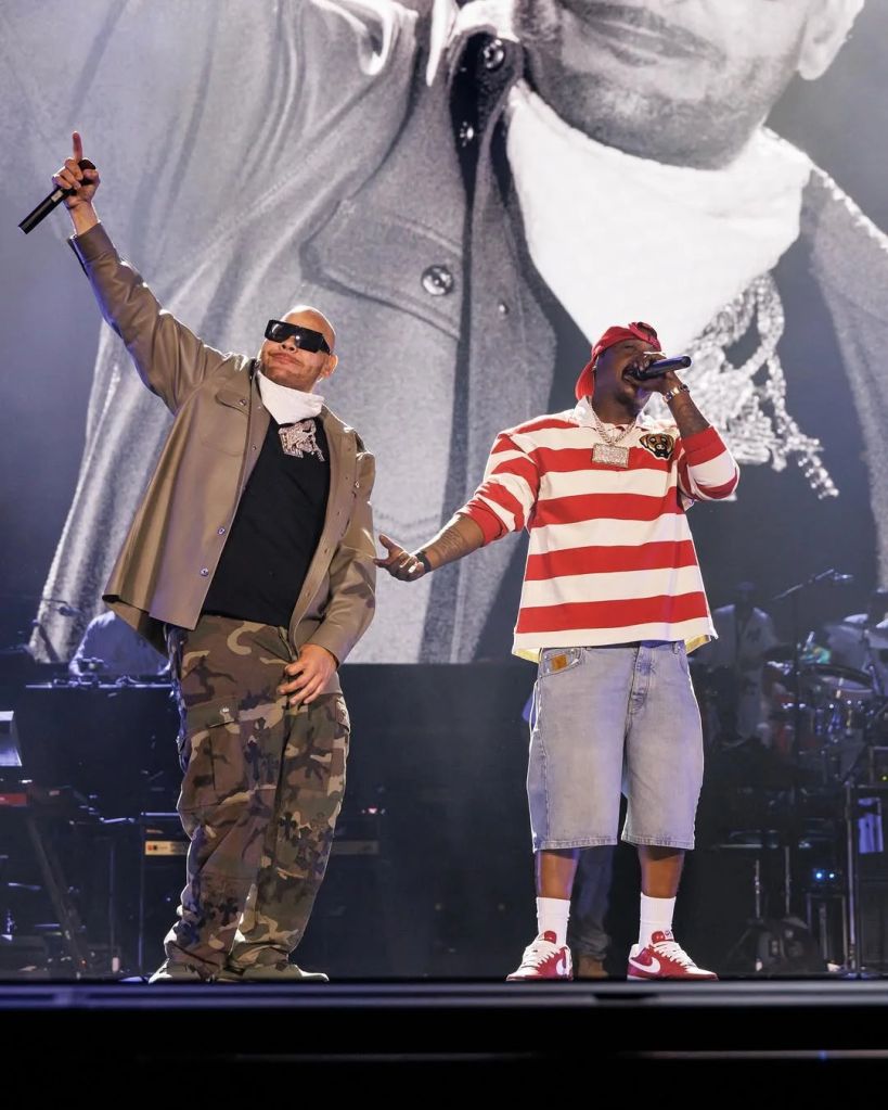 Two performers on stage, one wearing camouflage pants and a black shirt, the other wearing a red and white striped shirt, both holding microphones and singing.