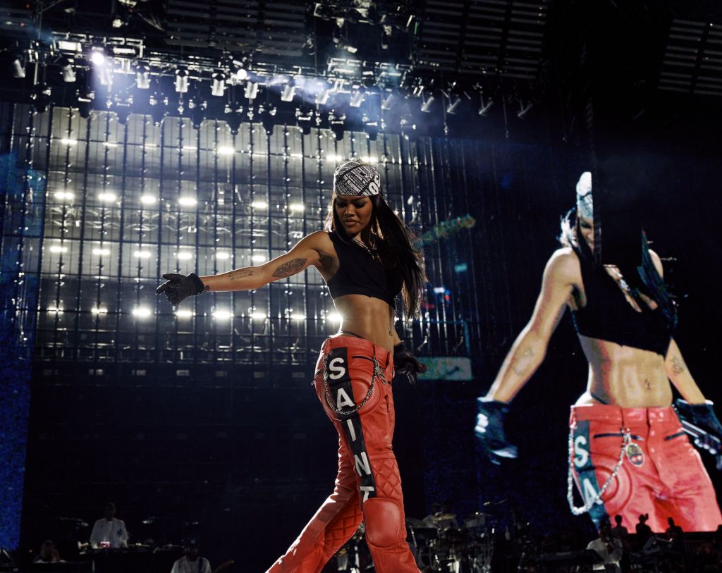 Performer on stage wearing black crop top, red pants with "SANA" text, and headpiece, dancing in front of bright lights and backdrop.